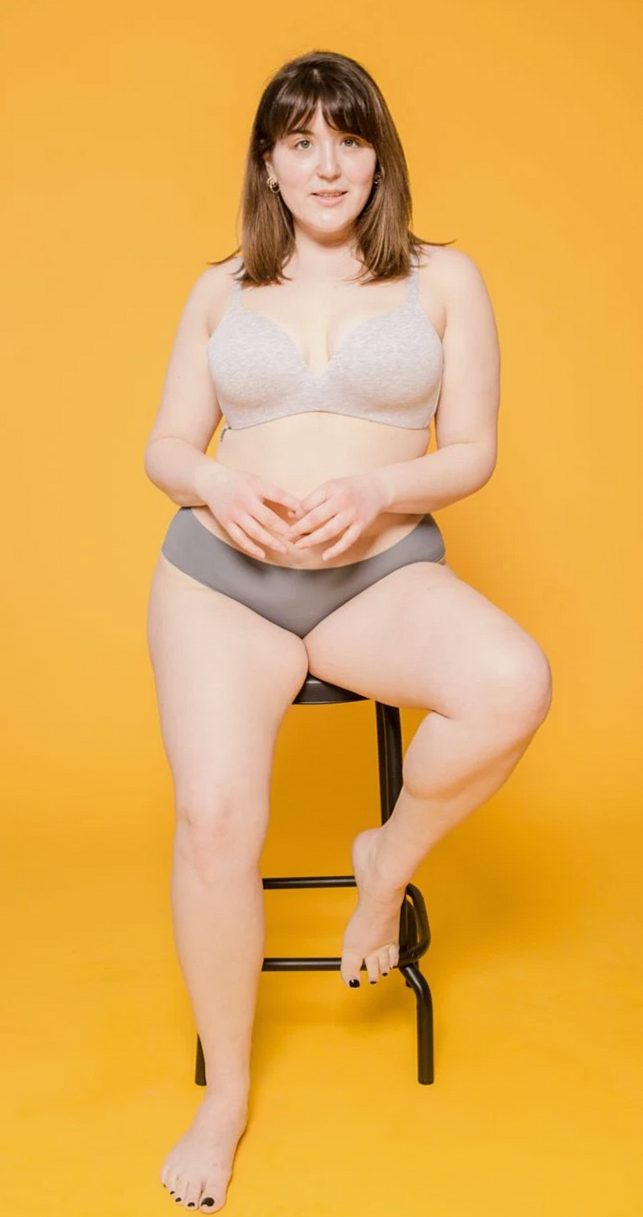 Plus size woman in bra and panties sitting on stool while looking at camera on yellow background